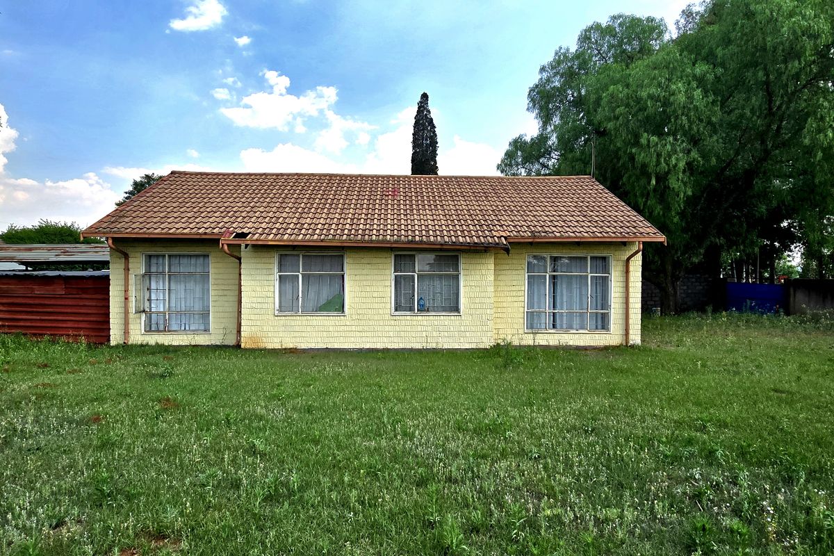 Rear view of a single-story brick house featuring a terracotta tile roof and large windows, set on a spacious, overgrown garden plot.