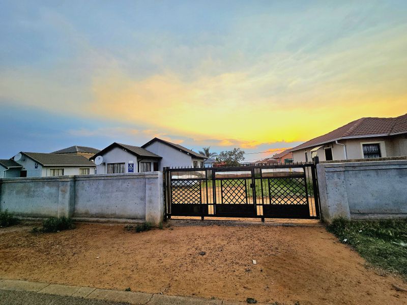 Front view of a residential property featuring a secure metal driveway gate and perimeter boundary wall under a sunset sky.