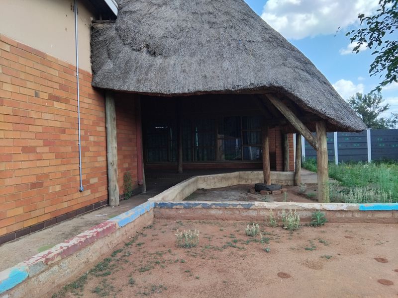 A covered patio area featuring a traditional thatched roof, exposed brickwork, and timber support posts, overlooking an outdoor space with a low concrete retaining wall.