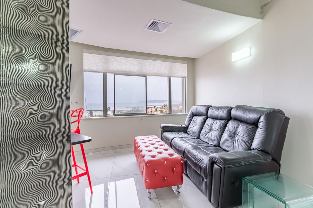 A bright living room featuring polished tiled flooring, a textured feature wall, and large sliding windows offering scenic coastal views.