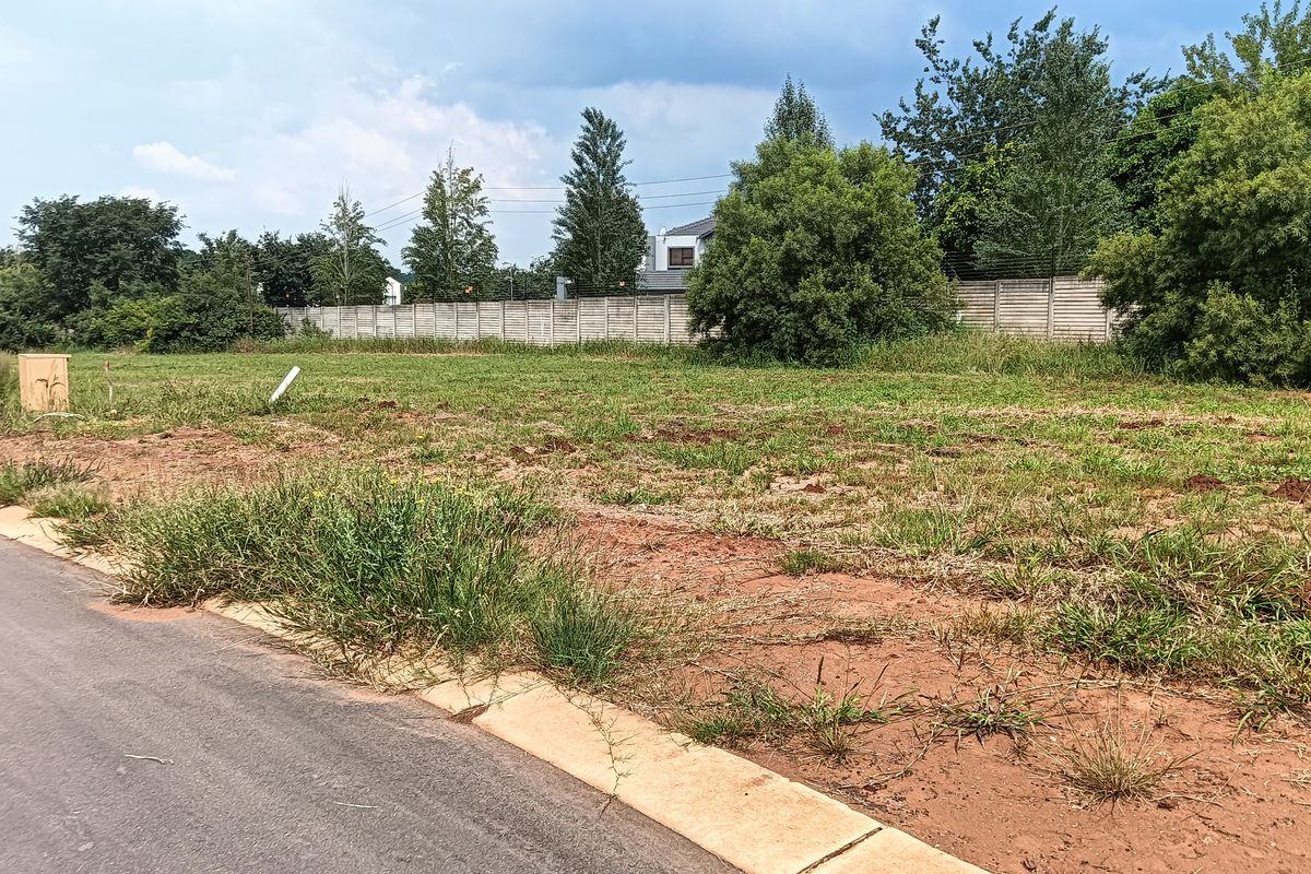 A vacant residential plot of land featuring a concrete boundary wall, accessible from a paved road with established curb infrastructure.