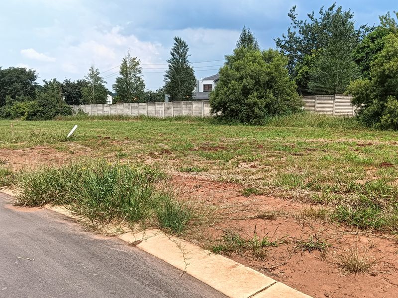 A vacant residential plot of land featuring a concrete boundary wall, accessible from a paved road with established curb infrastructure.