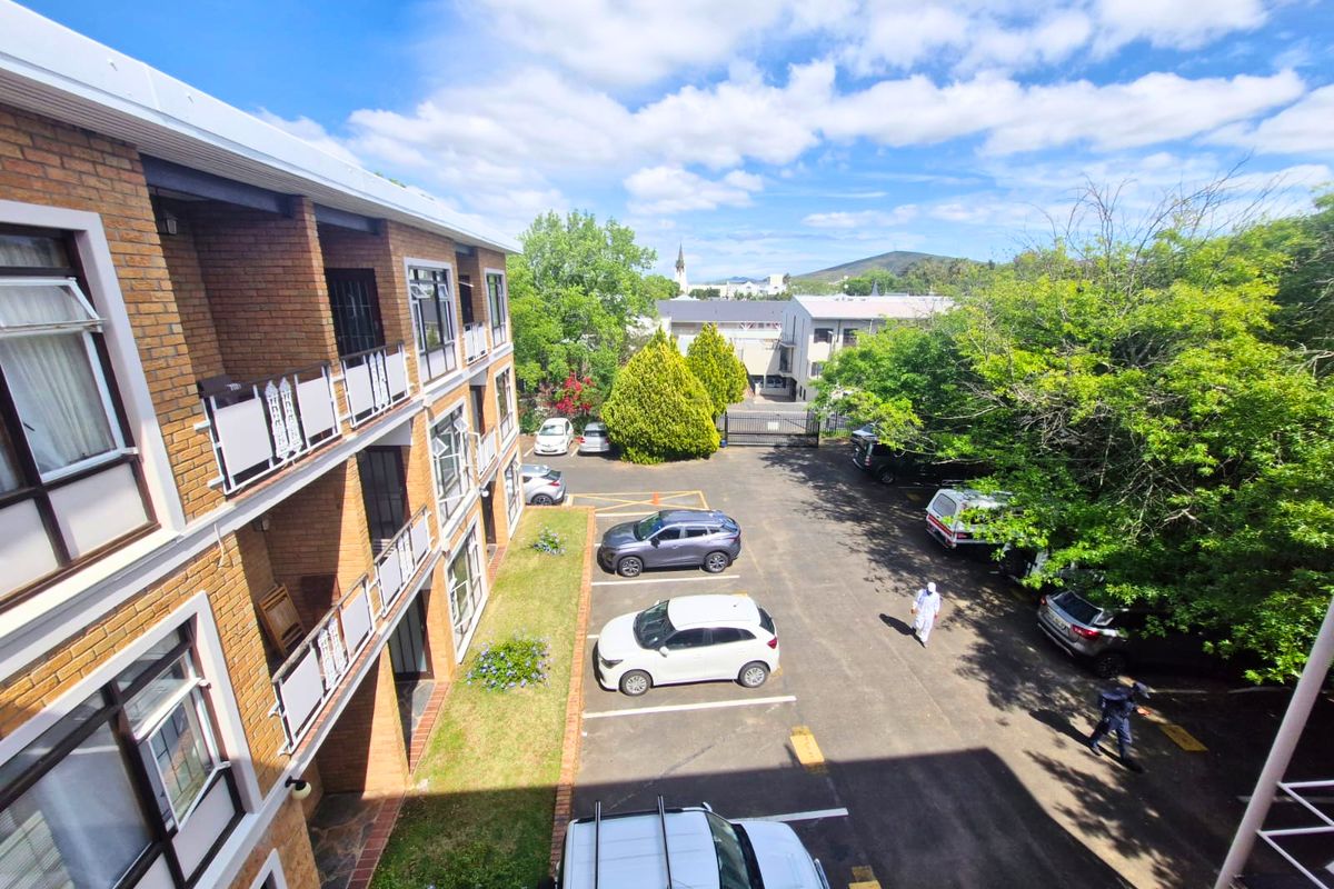 An elevated view of the apartment building's rear facade, featuring face brick construction, private balconies, and an adjacent paved parking area with landscaped greenery.