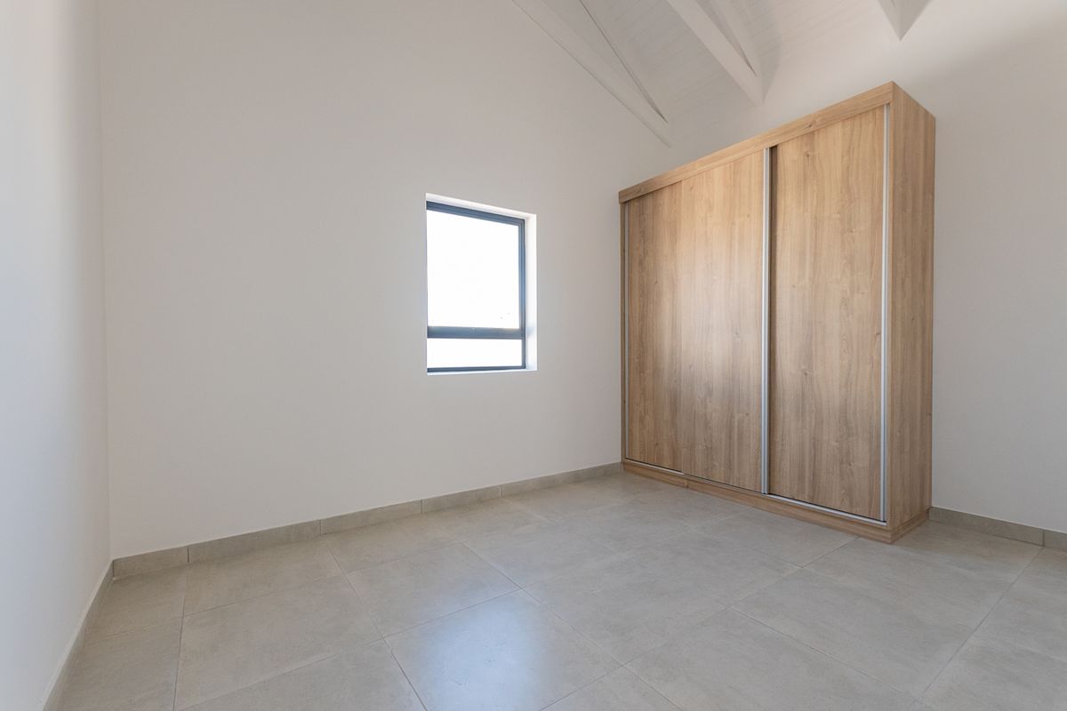 A bright, modern bedroom featuring neutral tiled flooring, a vaulted ceiling, and a large built-in wooden wardrobe.