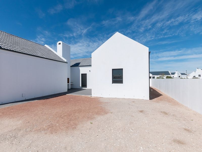Rear view of a modern white house featuring a tiled roof, chimney, built-in braai, and a paved patio area with an unlandscaped garden.
