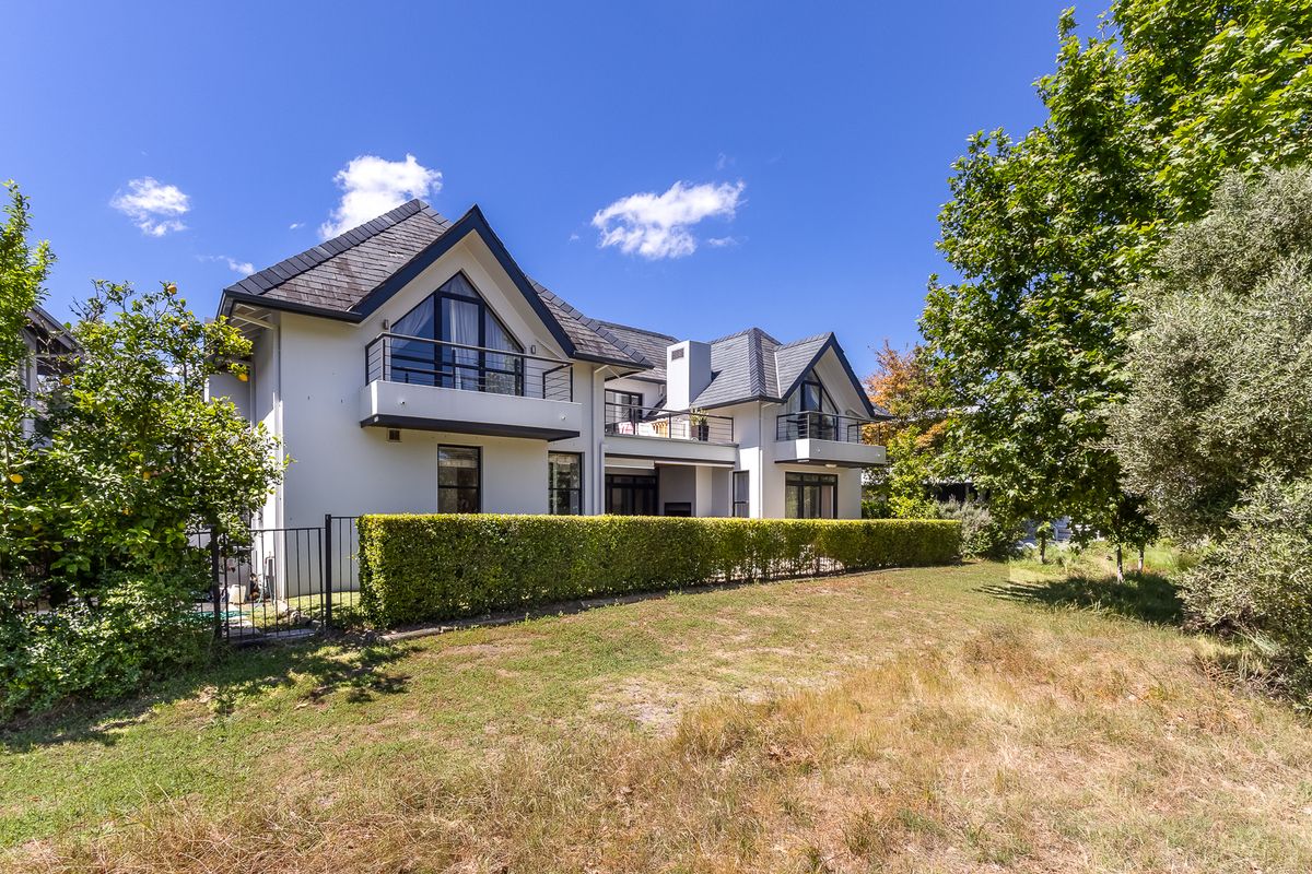Rear view of a modern multi-story residence featuring pitched tiled roofs, private balconies with metal railings, and a landscaped garden with established hedging.