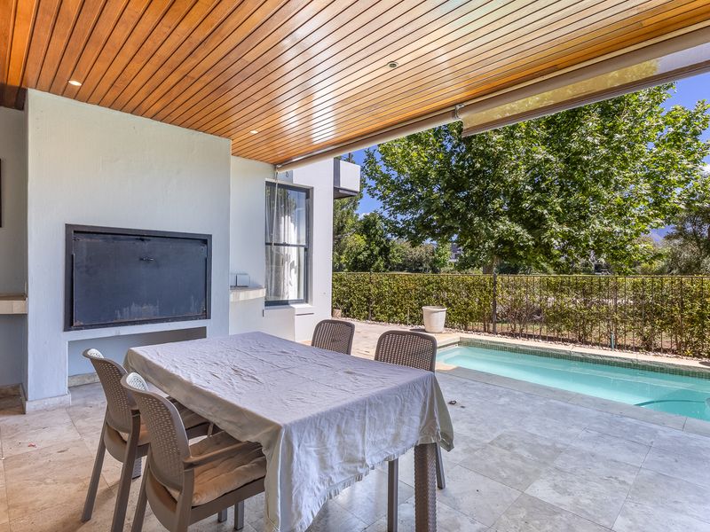 Covered outdoor patio featuring a timber-clad ceiling, built-in braai, and stone tile flooring, overlooking a private swimming pool and landscaped garden.