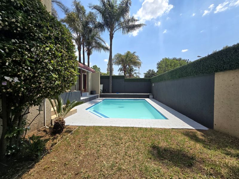A well-maintained outdoor pool area featuring a clear swimming pool, paved surround, and high boundary walls with privacy screening.