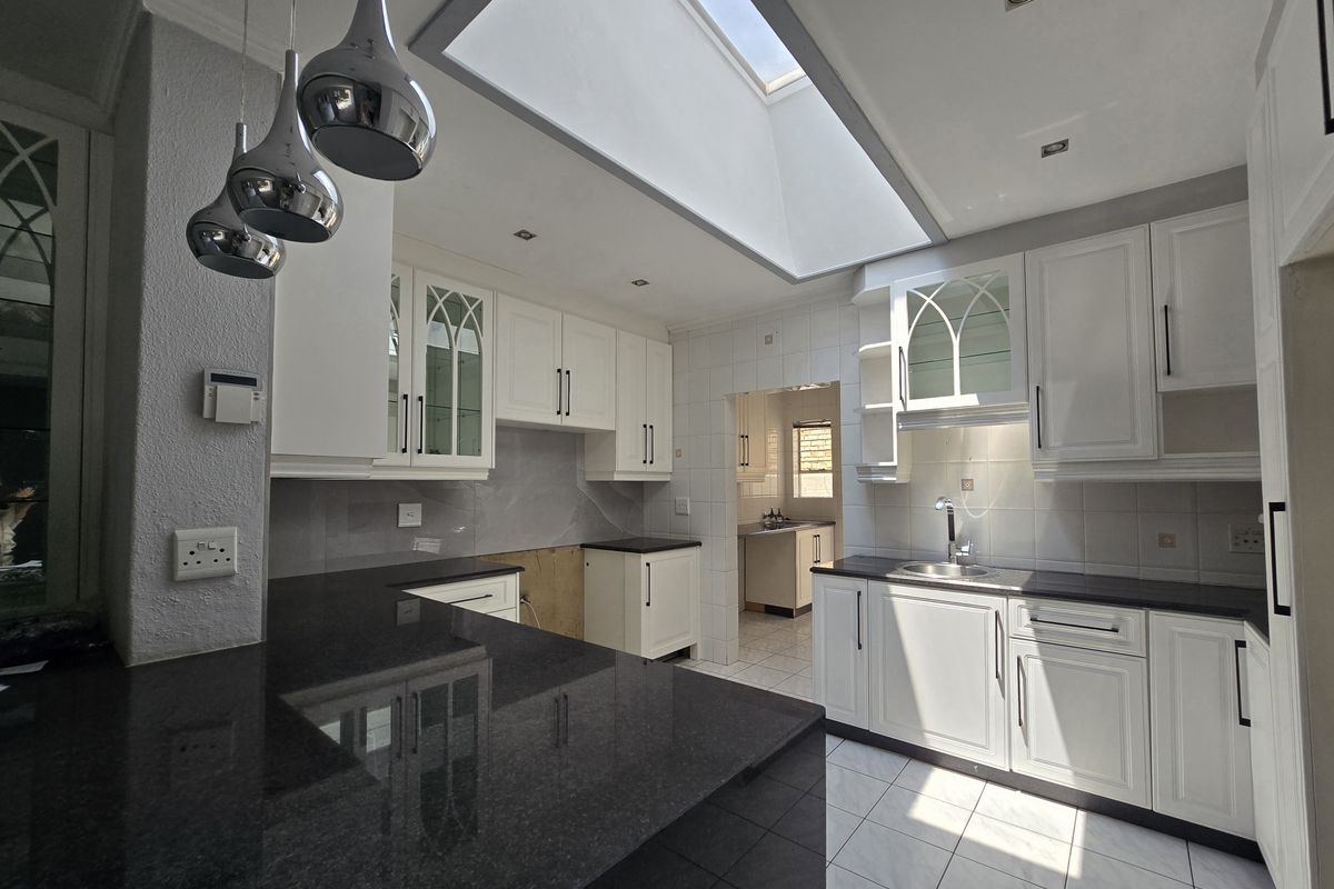 A bright, spacious kitchen featuring white cabinetry, dark granite countertops, a large skylight, and a connected scullery area, currently awaiting final finishing touches.