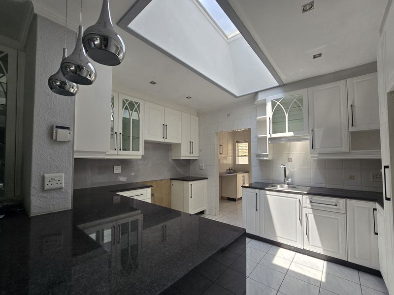 A bright, spacious kitchen featuring white cabinetry, dark granite countertops, a large skylight, and a connected scullery area, currently awaiting final finishing touches.