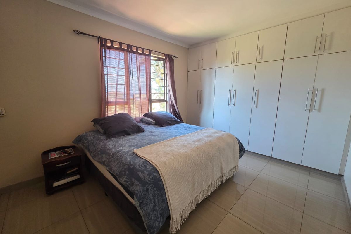A well-maintained bedroom featuring extensive floor-to-ceiling built-in cupboards, neutral tiled flooring, and a window fitted with security bars.