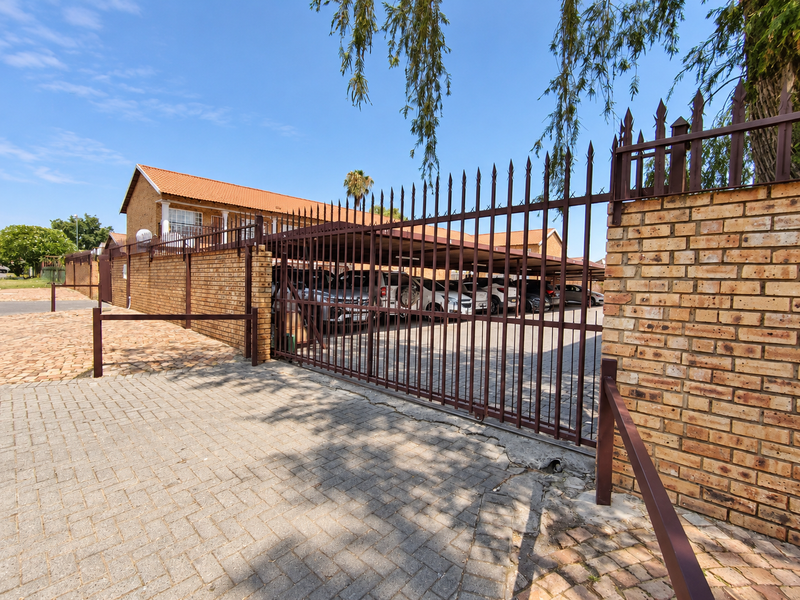 Exterior view of the apartment complex entrance featuring a secure metal gate, brick boundary walls, and a covered carport area for resident parking.