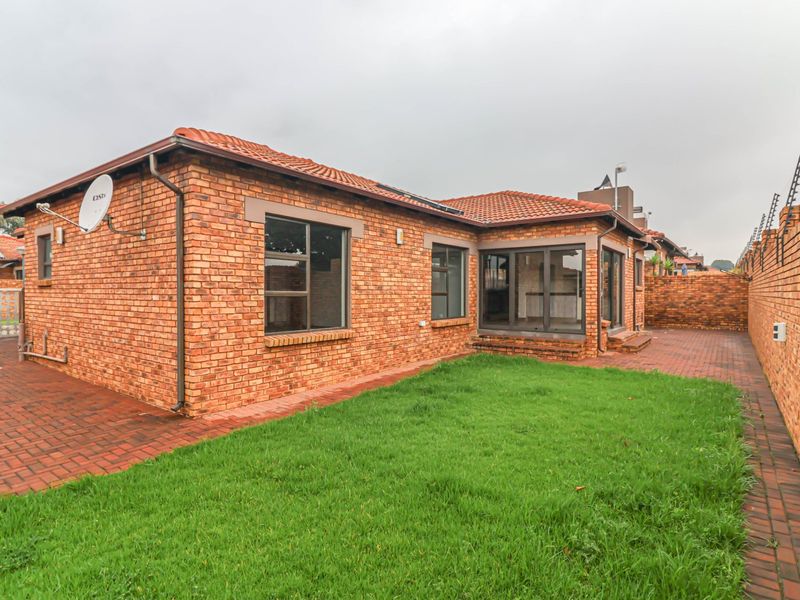 Rear view of a face brick cluster home featuring a paved patio area, small lawn, and secure boundary wall with electric fencing.