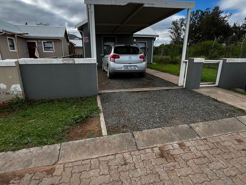 Front exterior view of the property featuring a gravel driveway, a covered carport, and a perimeter boundary wall.