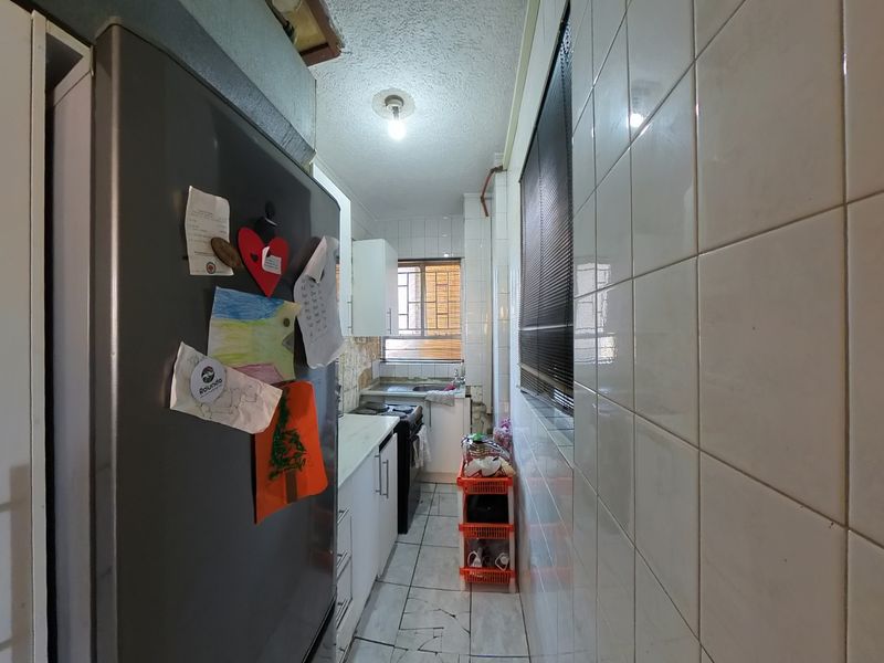 A narrow galley-style kitchen featuring tiled walls and flooring, built-in cabinetry, and a functional stove, requiring some cosmetic updates to the base cabinetry.