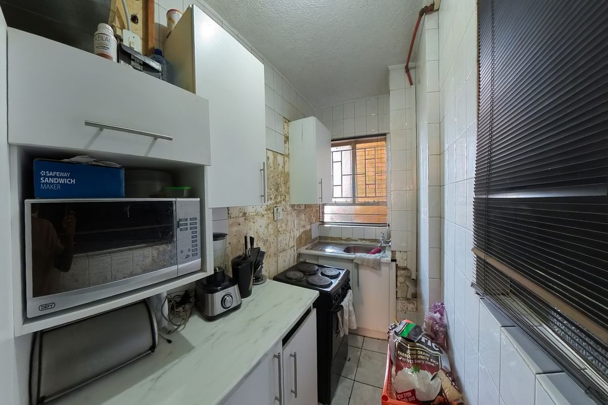 A compact kitchen featuring white cabinetry, a stainless steel sink, and an electric stove, currently requiring significant wall repairs and tile restoration.