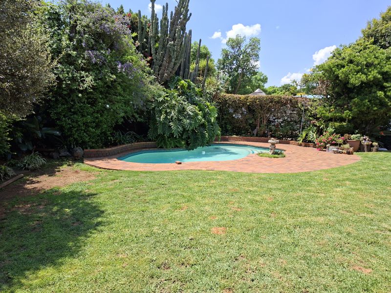 A private backyard featuring a swimming pool with a brick surround, set within a lush, mature garden with established trees and shrubs.