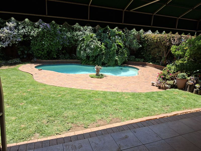 A well-maintained outdoor pool area featuring a curved swimming pool, brick paved surround, lush garden landscaping, and a covered tiled patio.