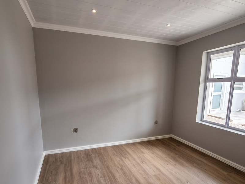 A bright, empty bedroom featuring modern timber-look laminate flooring, a clean tongue and groove ceiling with recessed lighting, and neutral grey walls.