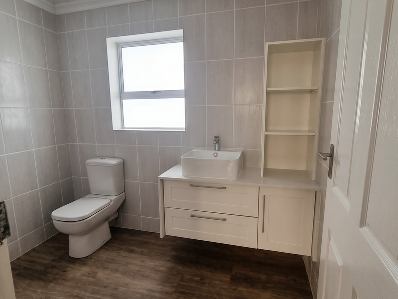 A clean, modern bathroom featuring a wall-mounted vanity with a vessel sink, built-in shelving, tiled walls, and wood-look flooring.