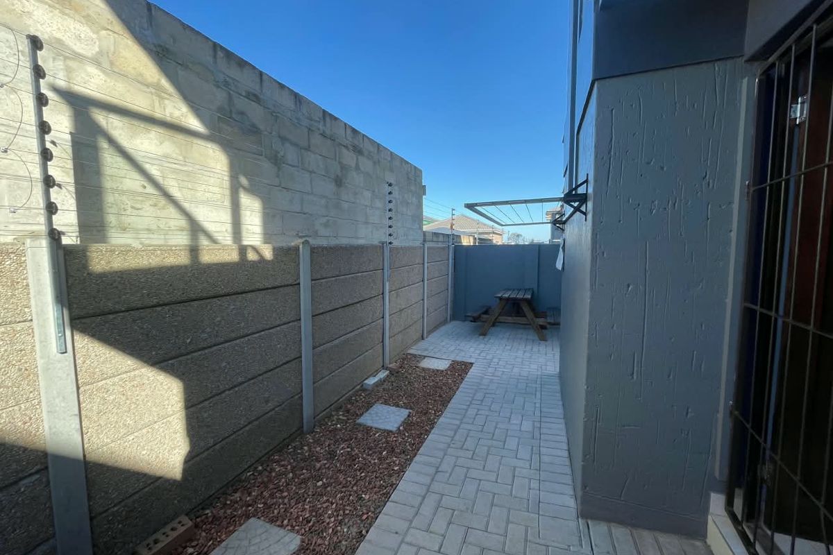 A side exterior area featuring a paved walkway, gravel garden bed, and secure precast concrete boundary wall with electric fencing.