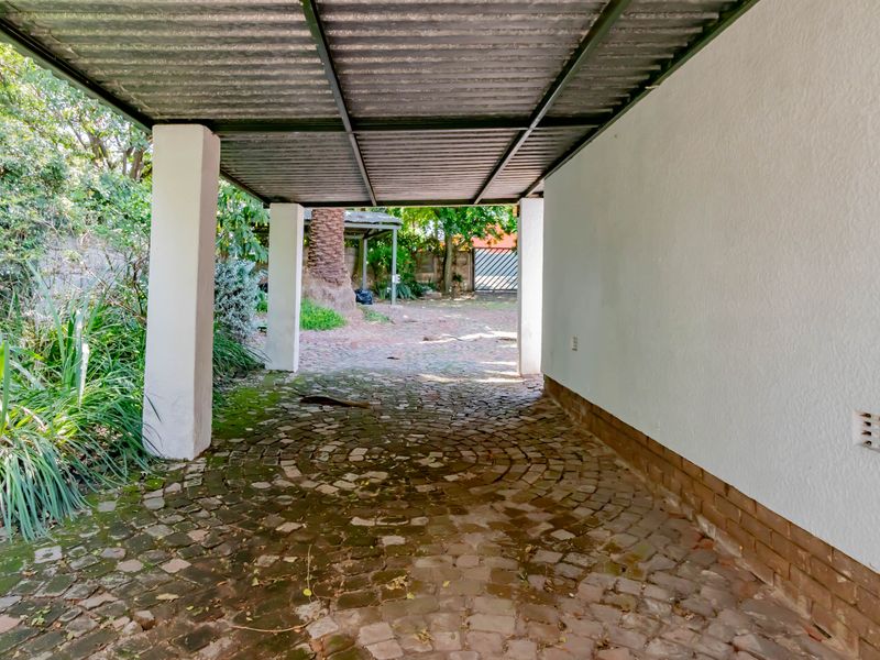 A covered carport area featuring a corrugated metal roof, concrete support pillars, and brick-paved flooring leading to the rear garden.