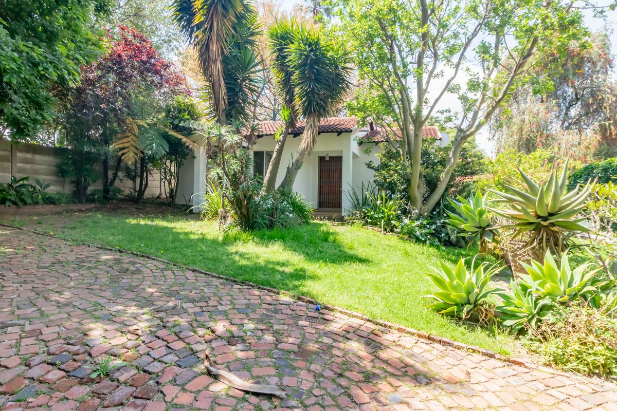 A view of the rear garden featuring a brick paved driveway, mature trees, established landscaping, and a white building with a tiled roof.