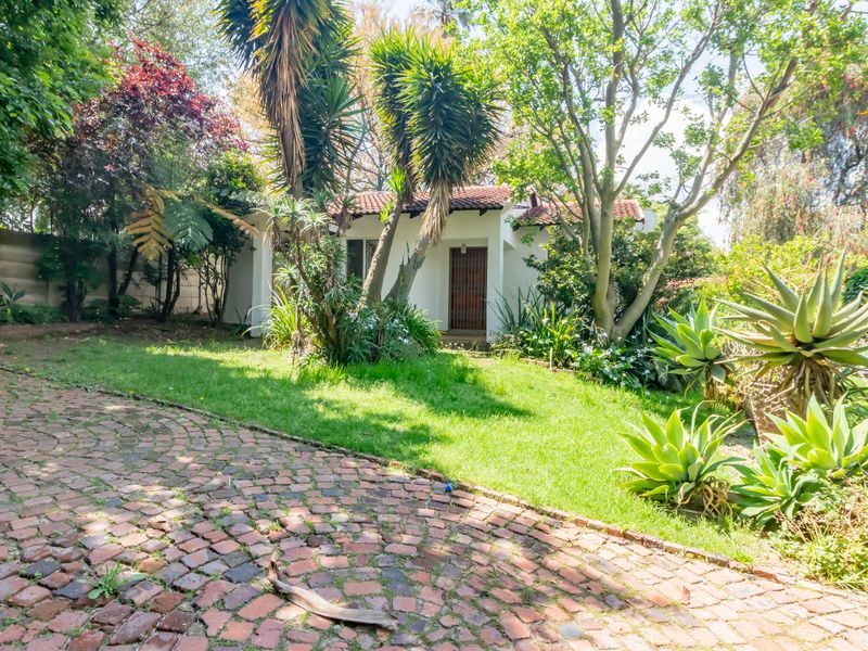 A view of the rear garden featuring a brick paved driveway, mature trees, established landscaping, and a white building with a tiled roof.