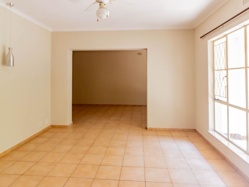 A bright, empty living area featuring neutral walls, tiled flooring, a ceiling fan, and large windows with security bars, leading into an adjacent room.