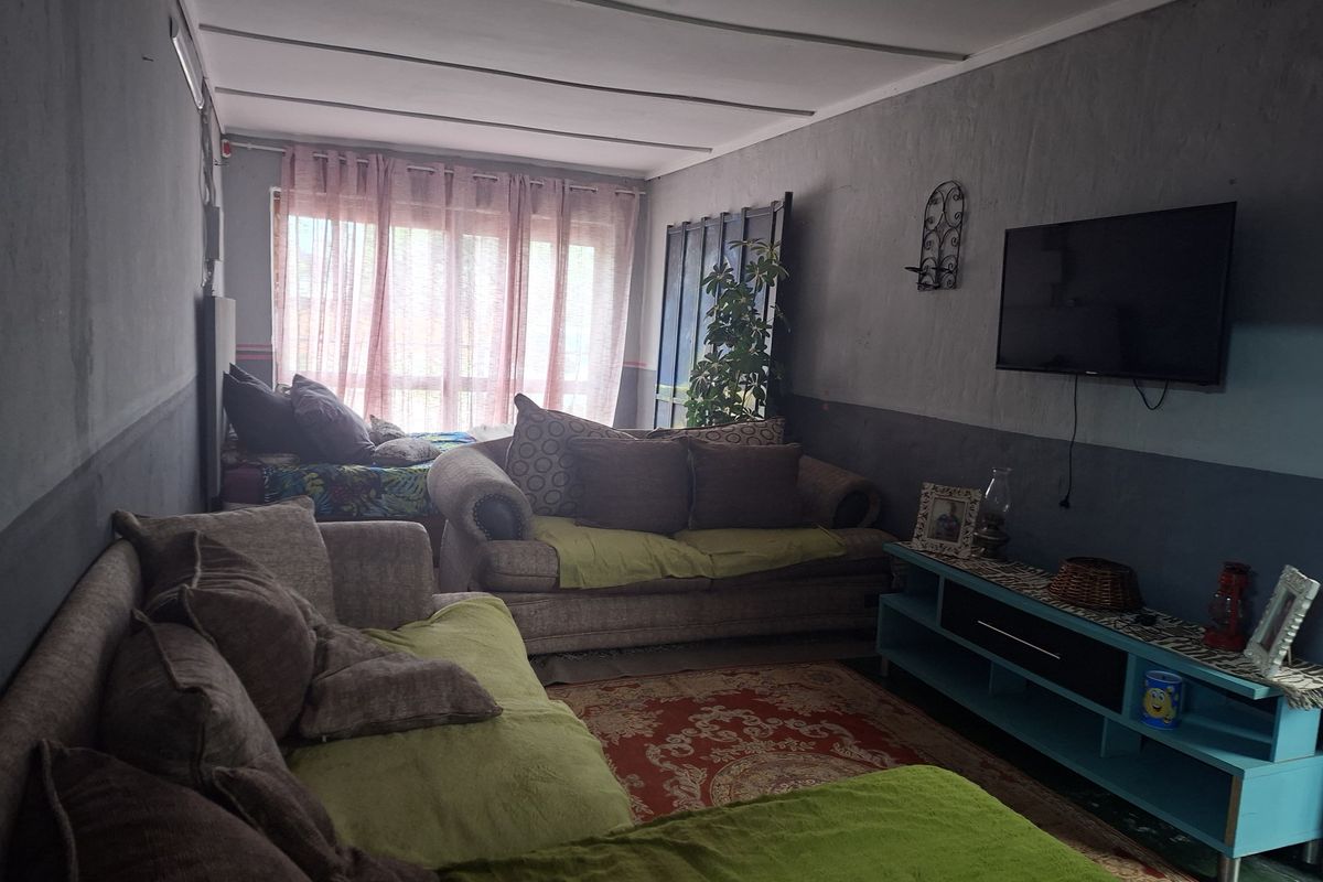 A living room space featuring a large window and panelled ceiling, currently used as a multi-functional living and sleeping area.