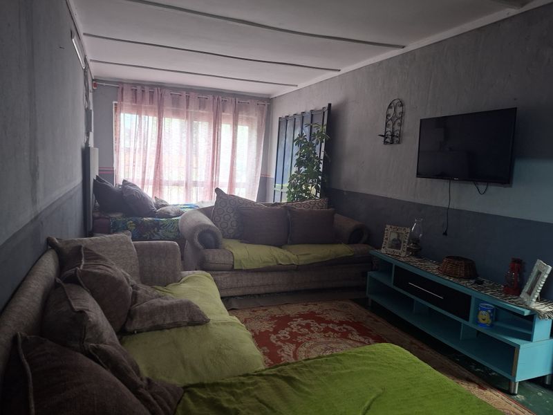 A living room space featuring a large window and panelled ceiling, currently used as a multi-functional living and sleeping area.