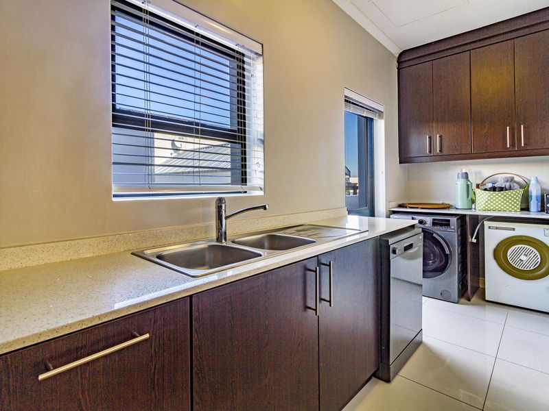 A functional laundry room featuring dark wood cabinetry, stone benchtops, a double stainless steel sink, and tiled flooring with space for under-counter appliances.