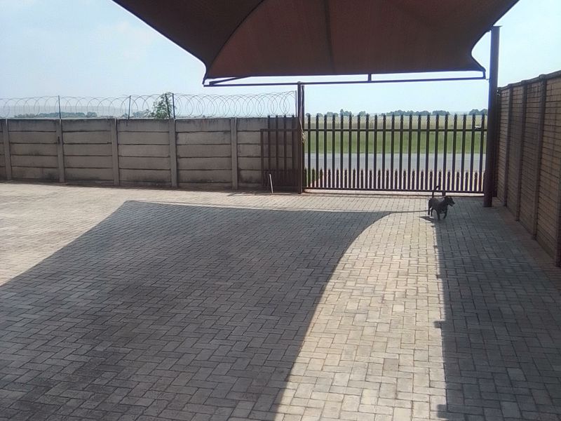 A paved outdoor parking area featuring a large shade sail, secure perimeter walling with razor wire, and a metal gate entrance.