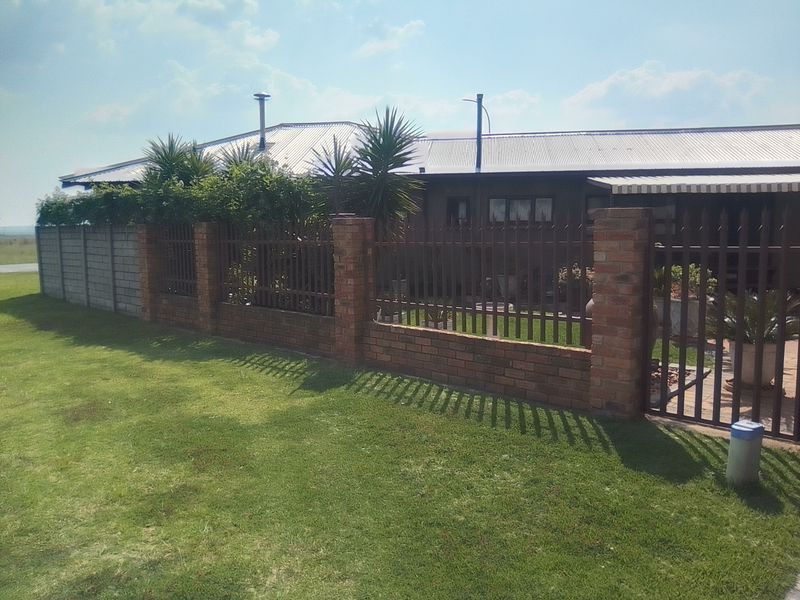 Front view of the property featuring a brick and metal palisade boundary fence, a well-maintained lawn, and a corrugated metal roof.
