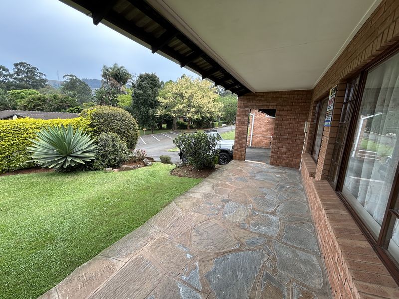 A covered patio area featuring natural stone paving and exposed brick walls, overlooking a well-maintained garden and driveway.