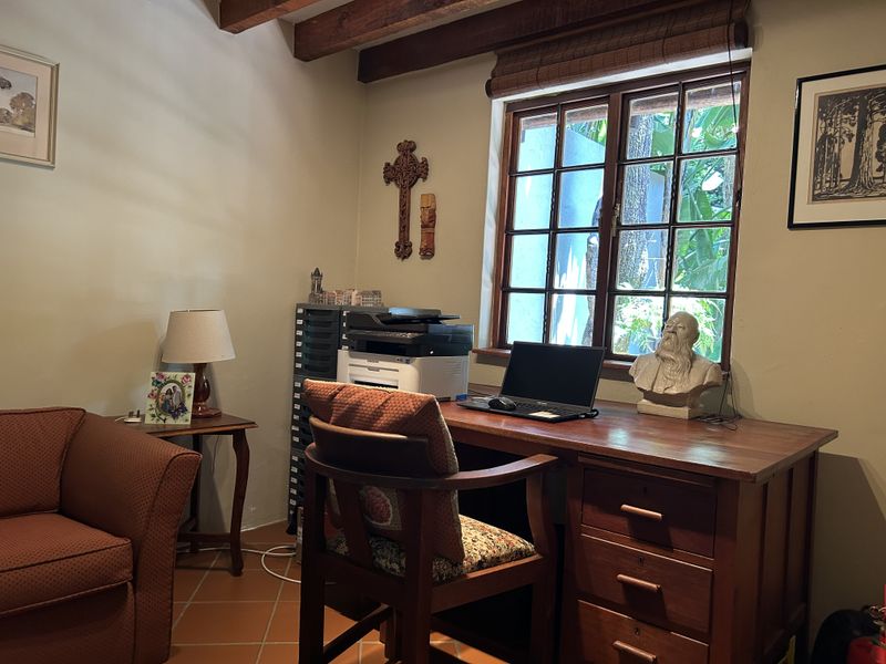 A cozy home office featuring exposed timber ceiling beams, terracotta floor tiles, and a wooden window frame overlooking the garden.