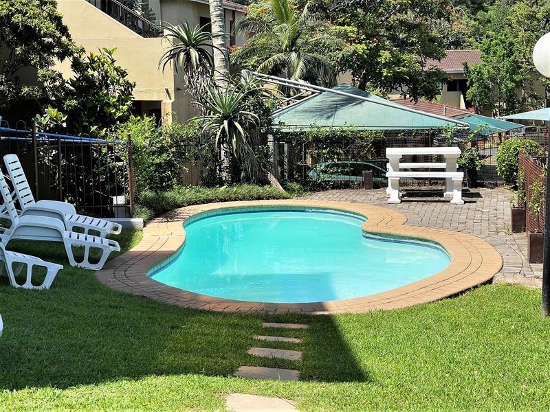 A private outdoor pool area featuring a kidney-shaped swimming pool, brick paved surround, and established garden lawn within a fenced complex setting.