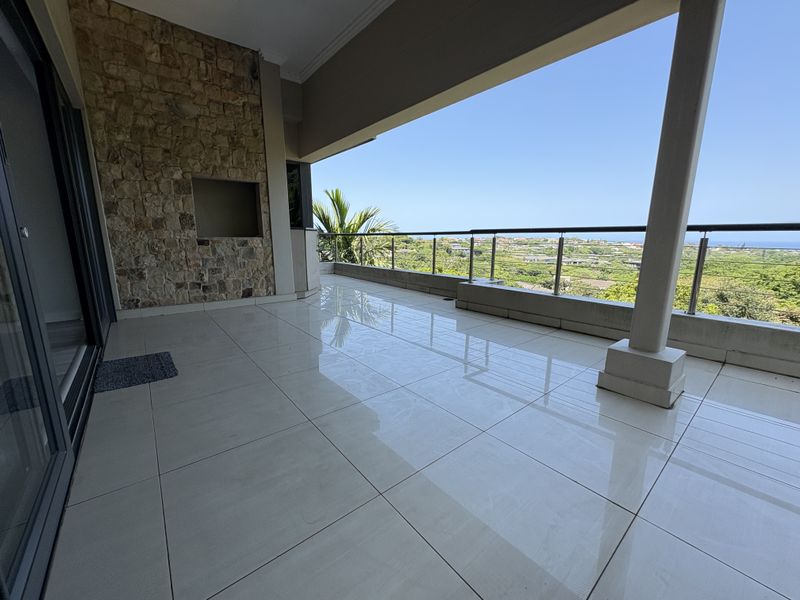 Spacious covered balcony featuring a built-in braai, stone cladding, and modern glass balustrades, offering expansive outdoor views.
