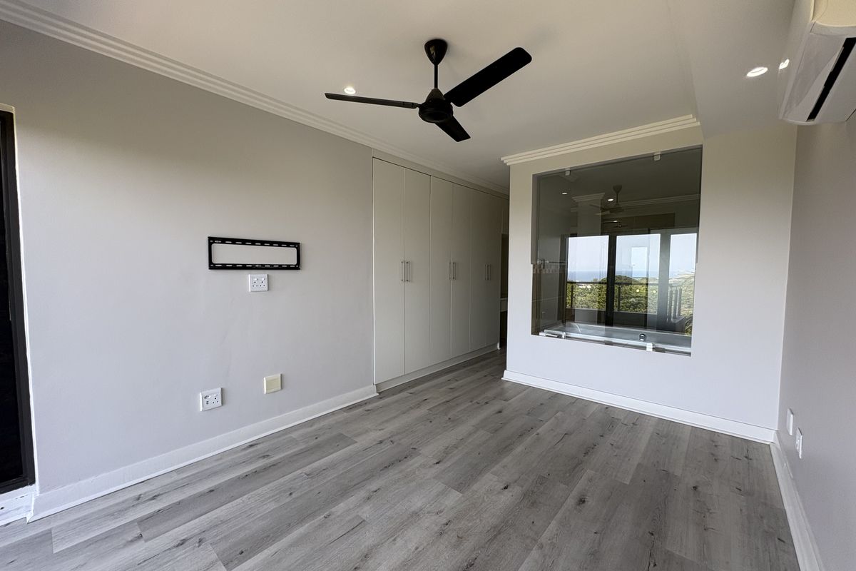 A bright, modern bedroom featuring light laminate flooring, built-in cupboards, a ceiling fan, and a unique glass partition wall looking into the en-suite bathroom.