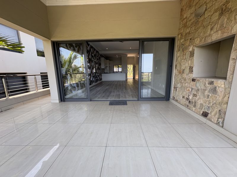 Covered patio featuring tiled flooring, a stone-clad built-in braai, and large sliding glass doors providing seamless access to the open-plan interior living space.