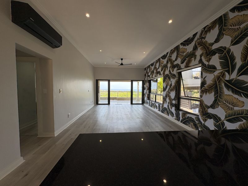 Spacious open-plan living area featuring modern laminate flooring, recessed lighting, a split-system air conditioner, and large sliding doors opening to a scenic balcony.