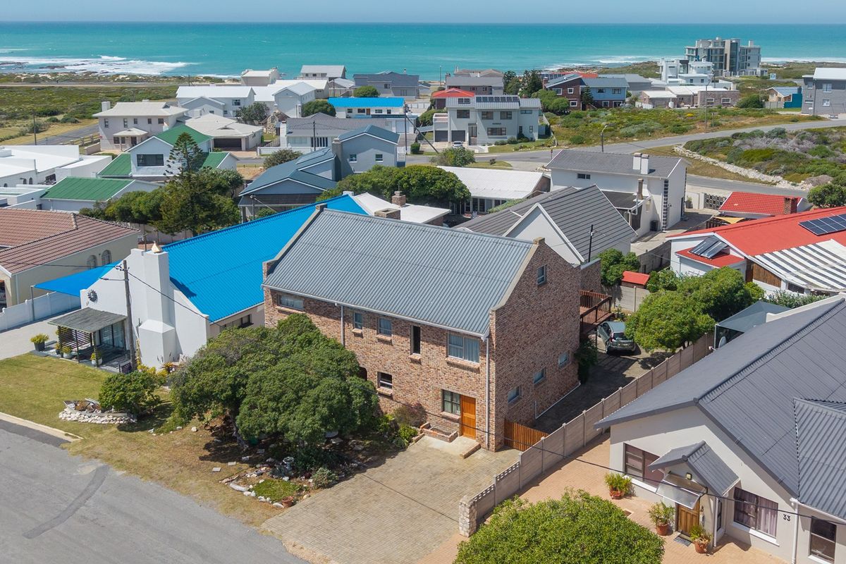 Aerial view of a multi-story brick house with a metal roof, featuring a paved driveway and surrounding residential neighborhood near the coast.