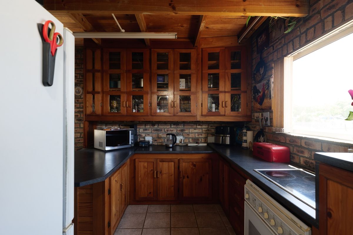 A rustic kitchen featuring exposed brick walls, timber cabinetry, exposed ceiling beams, and a functional layout with built-in oven and cooktop.