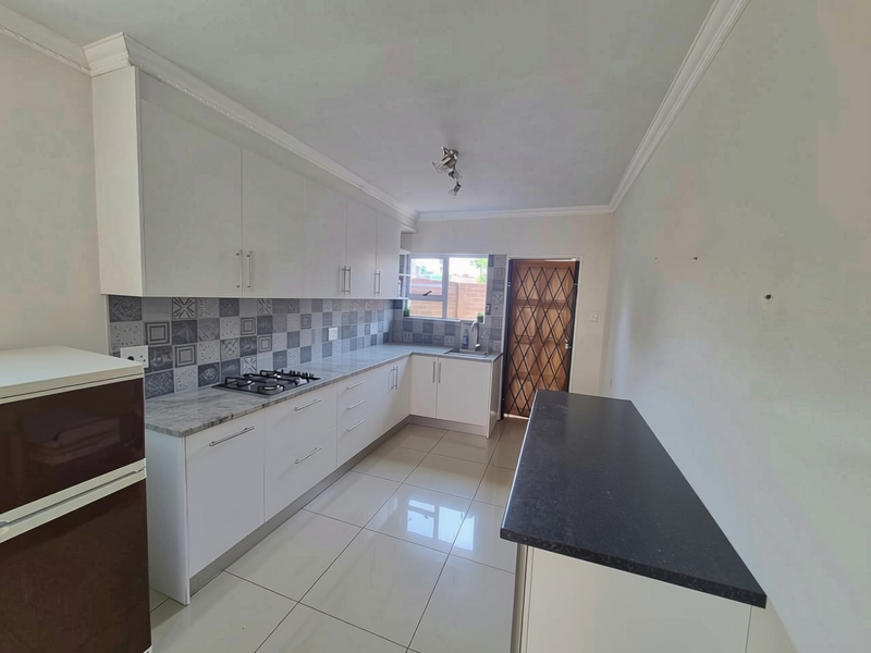 A clean, modern kitchen featuring white cabinetry, stone countertops, a gas hob, tiled backsplash, and polished floor tiles with direct access to the exterior.