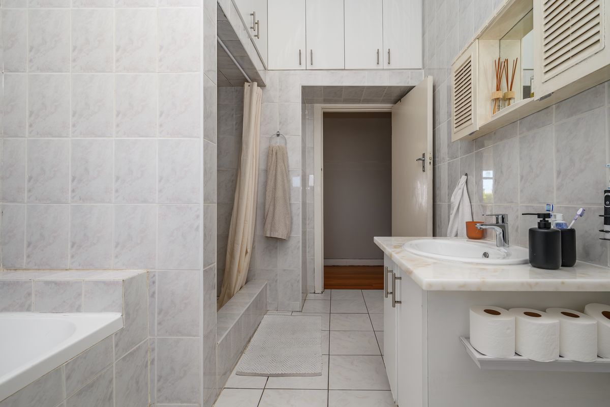 A functional bathroom featuring a built-in vanity with basin, overhead storage cupboards, a shower recess, and a bathtub, finished with neutral wall and floor tiles.