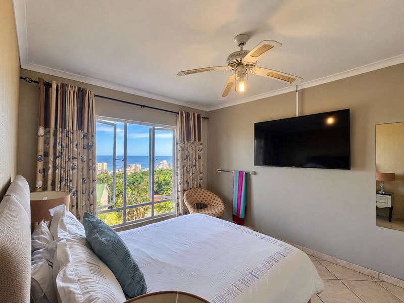 A well-maintained bedroom featuring tiled flooring, a ceiling fan, and a large window offering scenic coastal views.