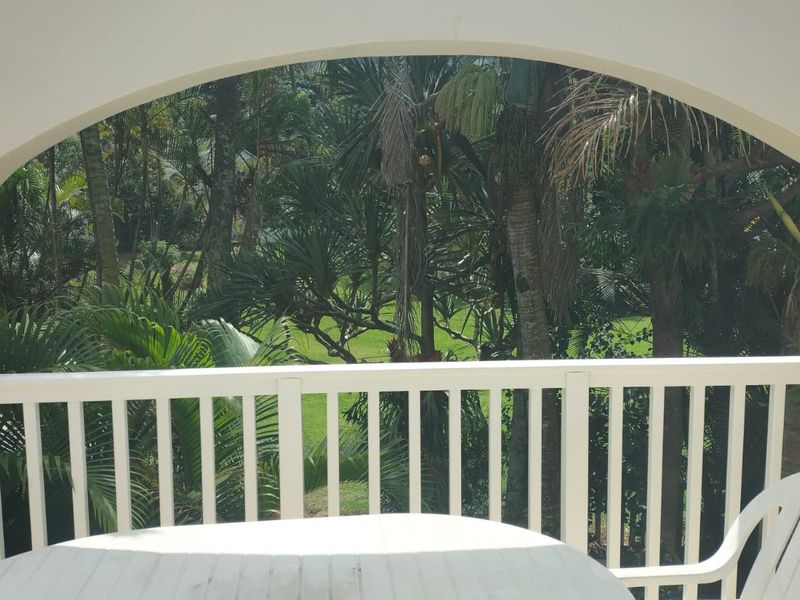 A covered balcony featuring an arched architectural opening and a white balustrade, overlooking a lush, green garden setting.