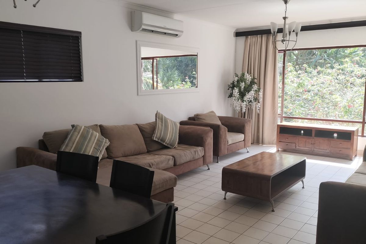 Spacious living area featuring tiled flooring, a wall-mounted air conditioning unit, and a large window providing natural light and garden views.