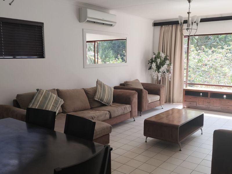 Spacious living area featuring tiled flooring, a wall-mounted air conditioning unit, and a large window providing natural light and garden views.
