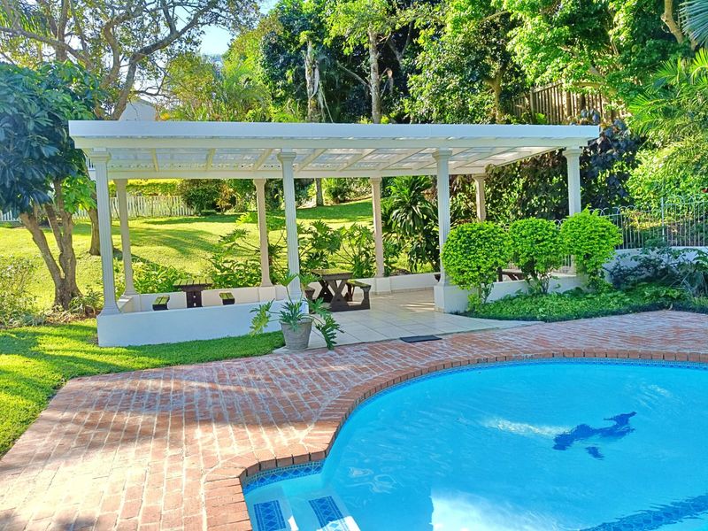 A well-maintained outdoor pool area featuring a brick-paved deck and a covered pergola, set within a lush, landscaped garden environment.